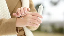 Trockene Haut: Eine Frau tr&auml;gt einen Wintermantel. Sie h&auml;lt eine wei&szlig;e Creme in der Hand. Mit der einen Hand tr&auml;gt sie die feuchtigkeitsspendende Creme auf die andere Hand auf.