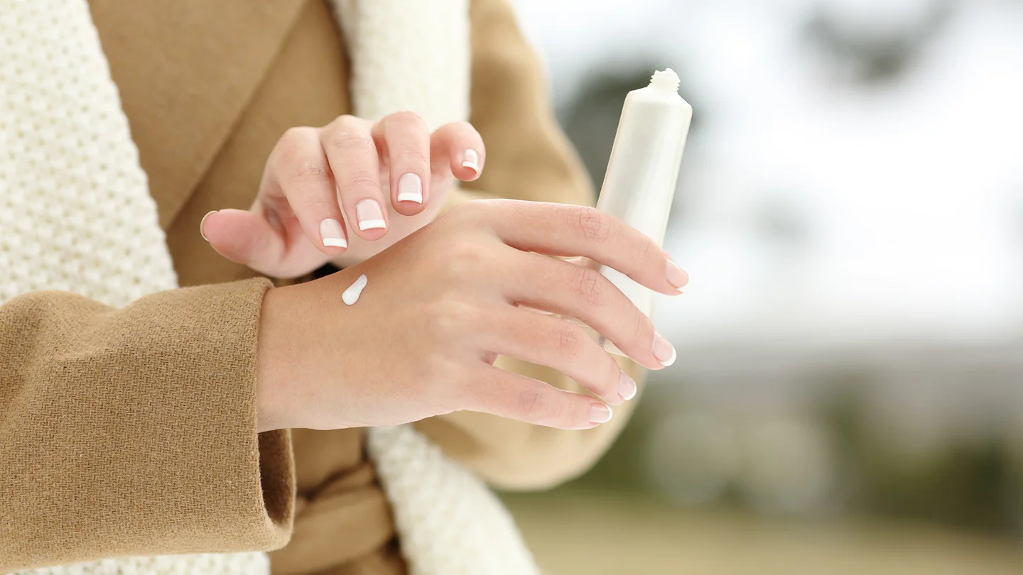 Trockene Haut: Eine Frau tr&auml;gt einen Wintermantel. Sie h&auml;lt eine wei&szlig;e Creme in der Hand. Mit der einen Hand tr&auml;gt sie die feuchtigkeitsspendende Creme auf die andere Hand auf.