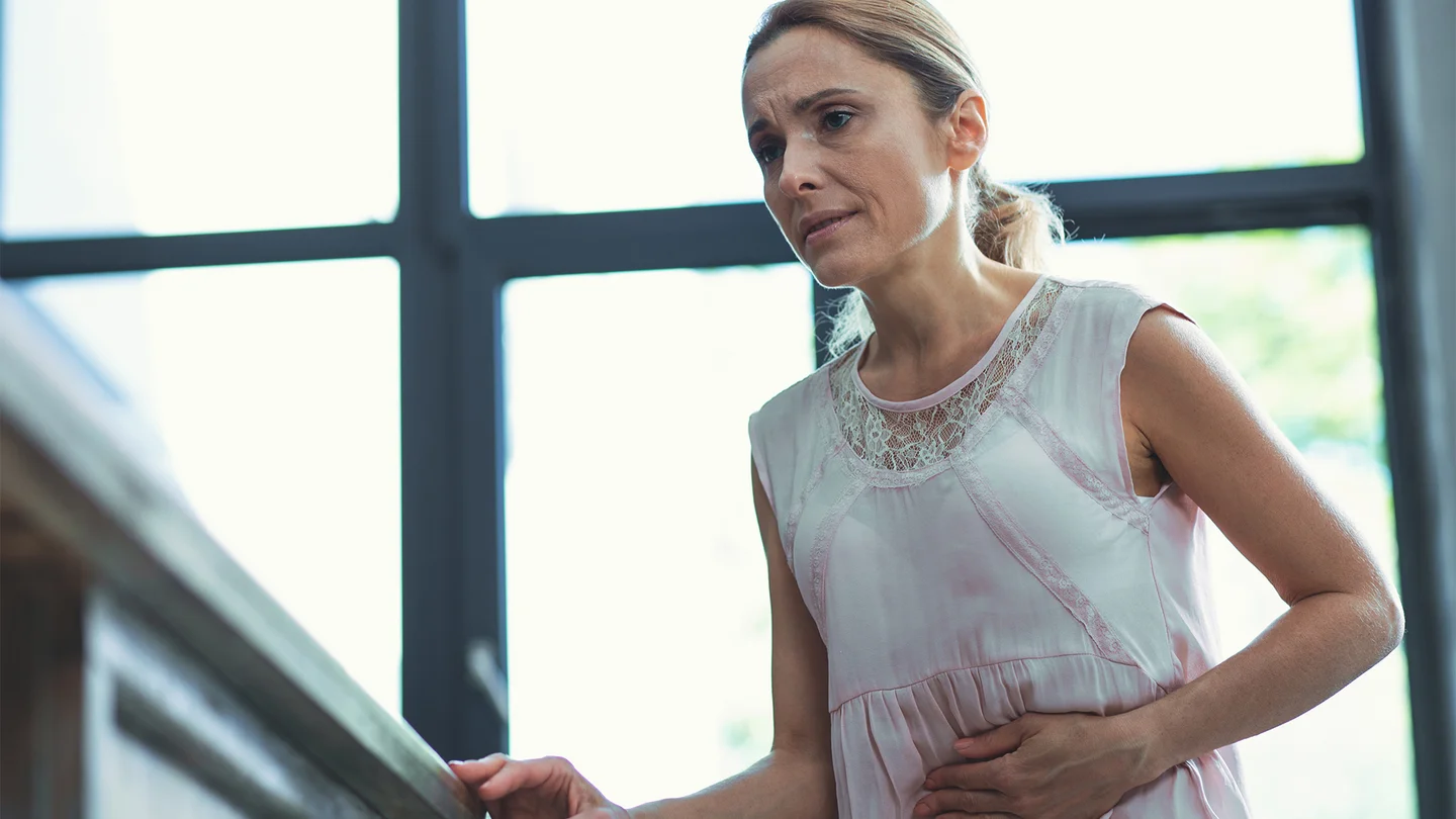 Magengeschwür: Eine Frau steht in ihrer Wohnung und hält sich den Bauch. Sie wirkt angespannt und als hätte sie Schmerzen.