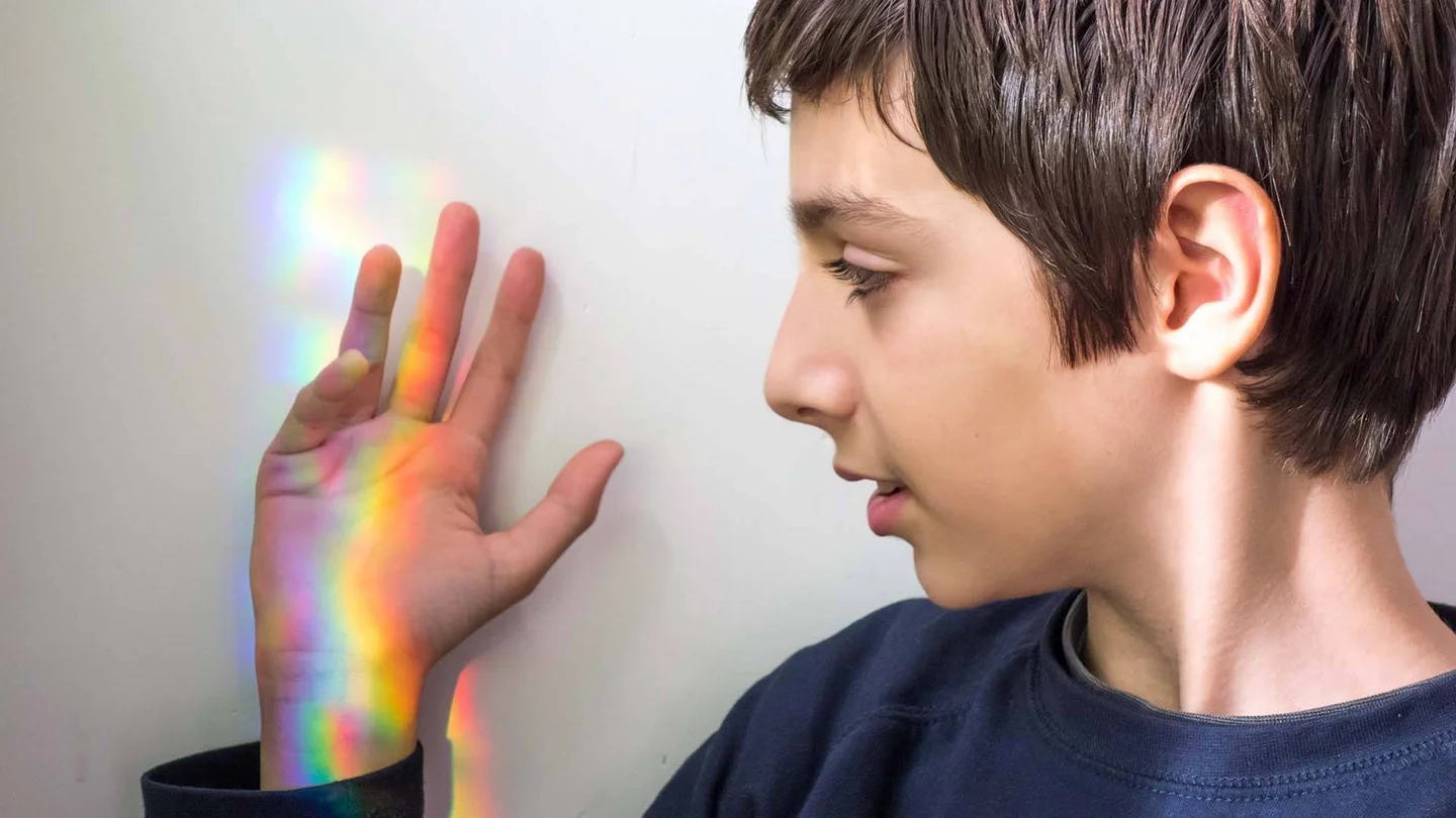 Ein Junge h&auml;lt seine Hand an eine wei&szlig;e Wand. Auf der Hand spiegeln sich die Regenbogenfarben von gebrochenem Sonnenlicht.
