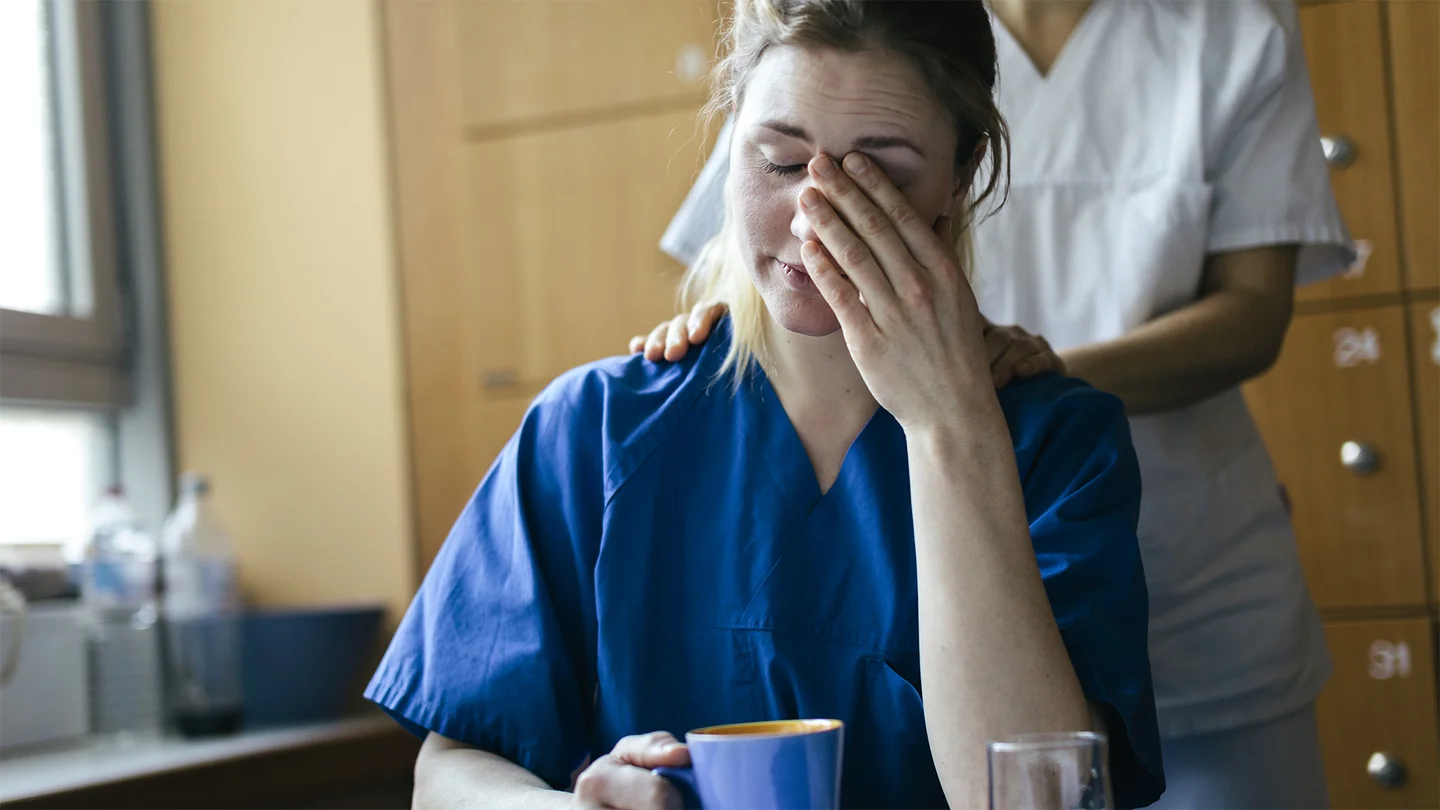 Eine Krankenpflegerin sitzt mit einer Tasse an einem Tisch. Mit dem linken Ellenbogen st&uuml;tzt sie sich auf und reibt mit ihrer Hand &uuml;ber ihr Gesicht. Eine Kollegin legt ihr von hinten tr&ouml;stend die H&auml;nde auf die Schulter.
