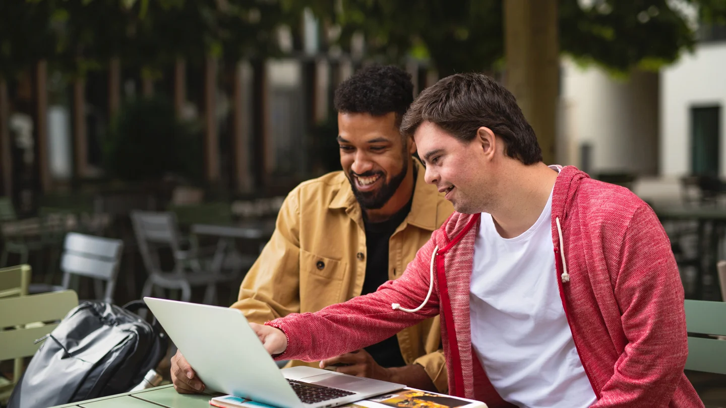 Ein junger Mann mit Behinderung und ein weiterer junger Mann schauen gemeinsam auf einen Laptop.