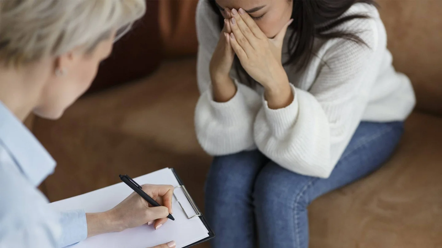 Eine junge Frau sitzt auf einer Couch, lehnt sich nach vorne und stützt verzweifelt den Kopf in ihre Hände. Ihr gegenüber sitzt eine Therapeutin, die sich Notizen macht.