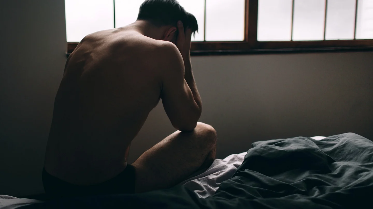 Ein junger Mann sitzt auf dem Bett und hält seinen gesenkten Kopf mit beiden Händen.