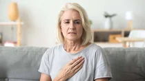 Eine Frau mittleren Alters im T-Shirt. Sie hat sich besorgt eine Hand auf den oberen Brustbereich gelegt.