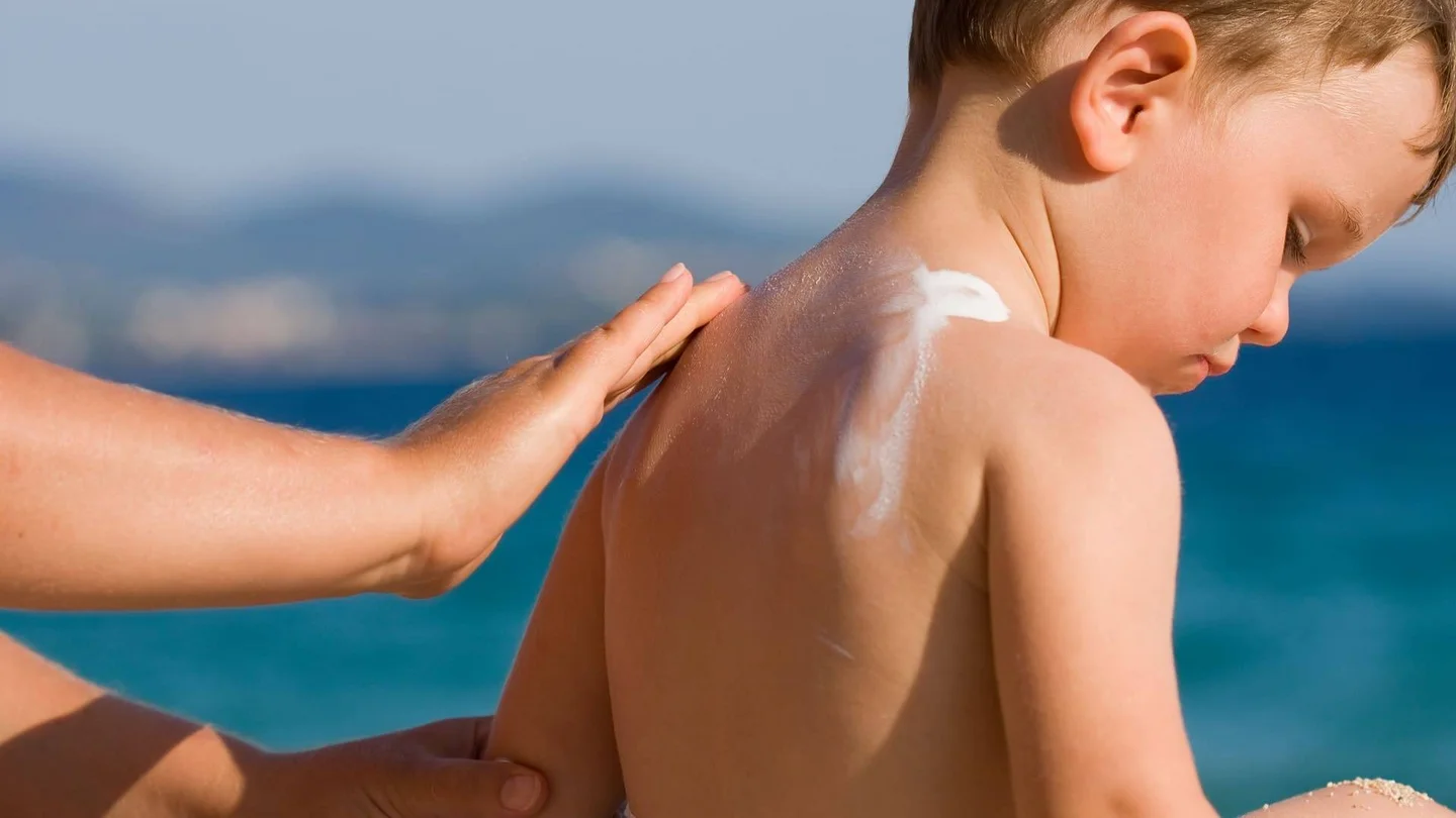 Ein Kleinkind sitzt am Strand und wird am Rücken eingecremt.