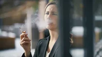 Eine Frau steht an einem Fenster, h&auml;lt eine Zigarette in der Hand und atmet Rauch aus.