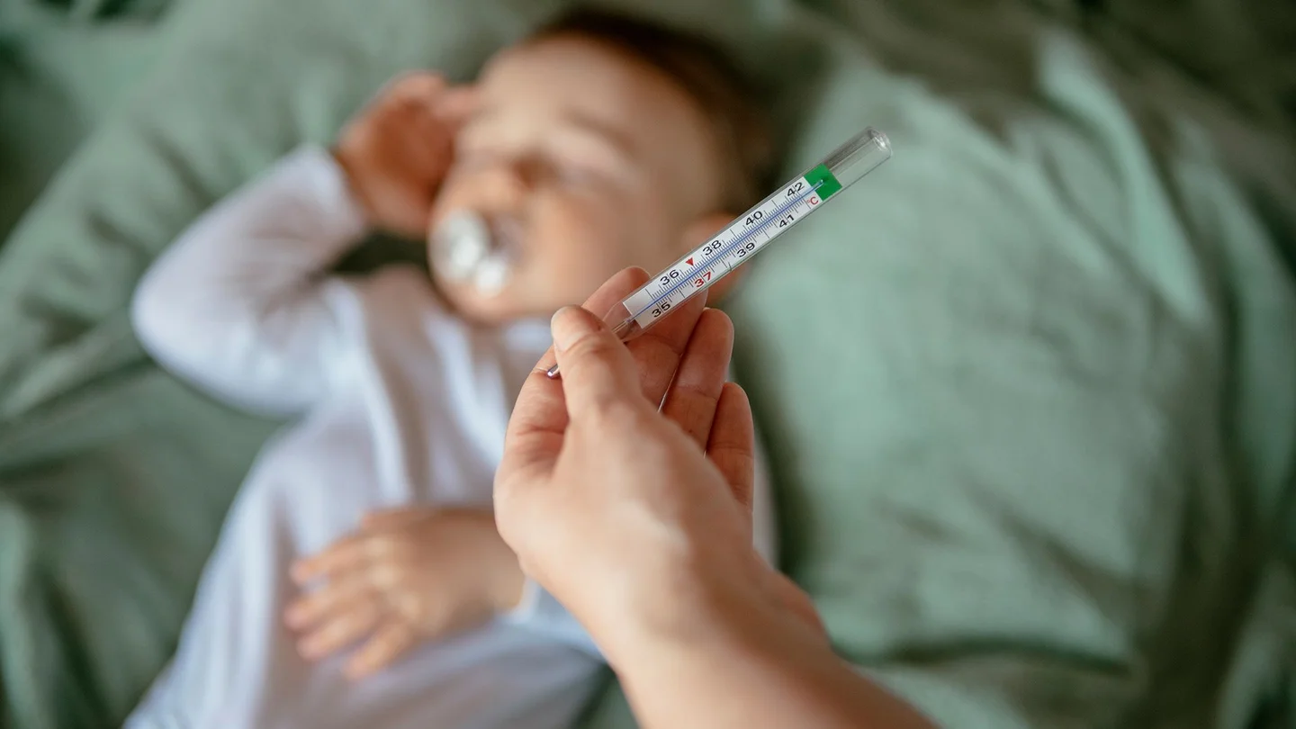 Eine Hand h&auml;lt ein Fieberthermometer &uuml;ber ein Baby