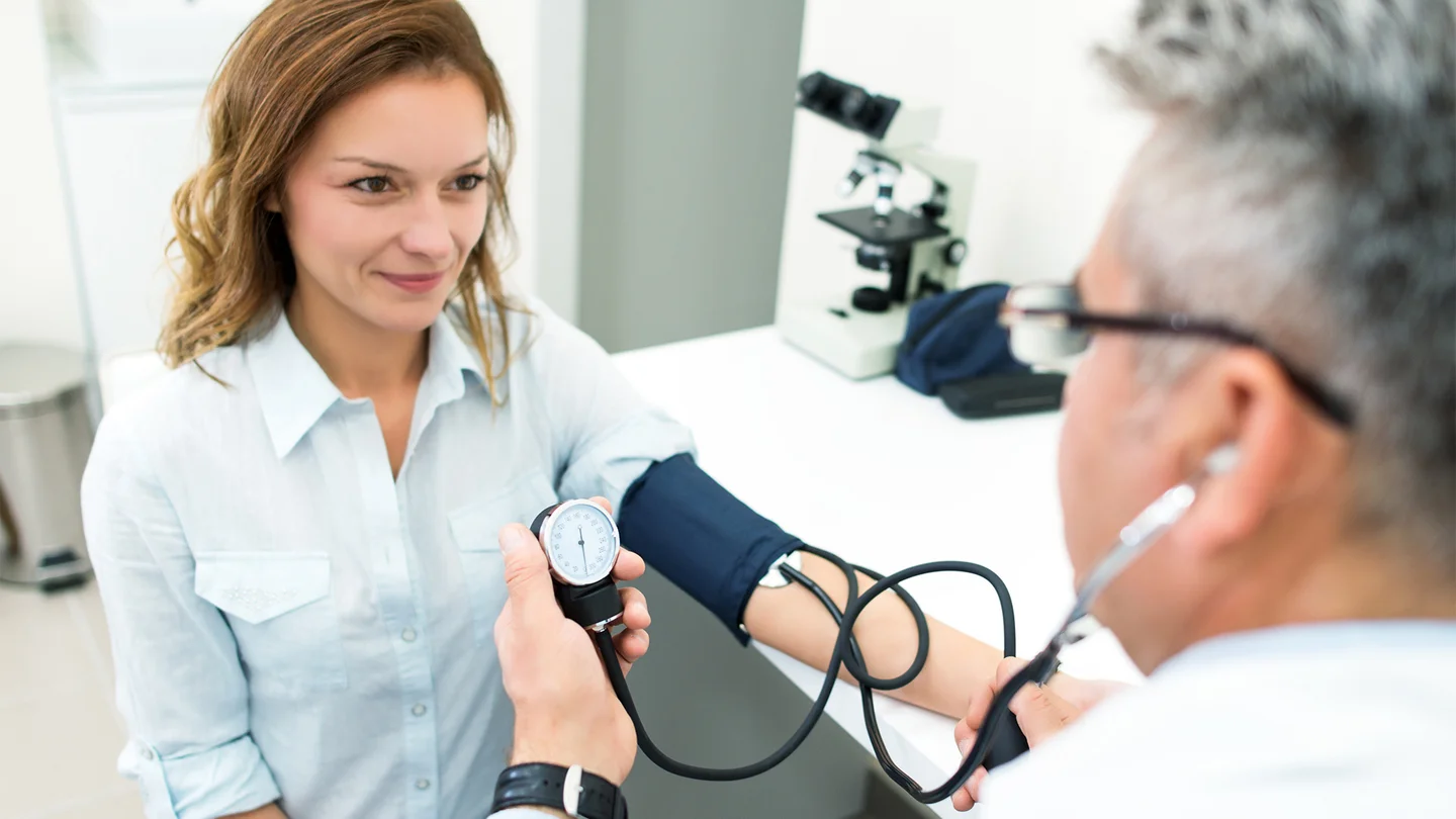 Gesundheits-Check-Up: Eine Frau sitzt einem Arzt gegen&uuml;ber. Sie tr&auml;gt eine Manschette um den Arm, ihr Blutdruck wird gemessen.