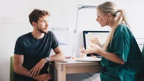 Hodenkrebs: Ein Mann sitzt in einem Behandlungszimmer. Sein Blick ist auf eine &Auml;rztin gerichtet, die ihm gegen&uuml;ber sitzt und mit ihm spricht.