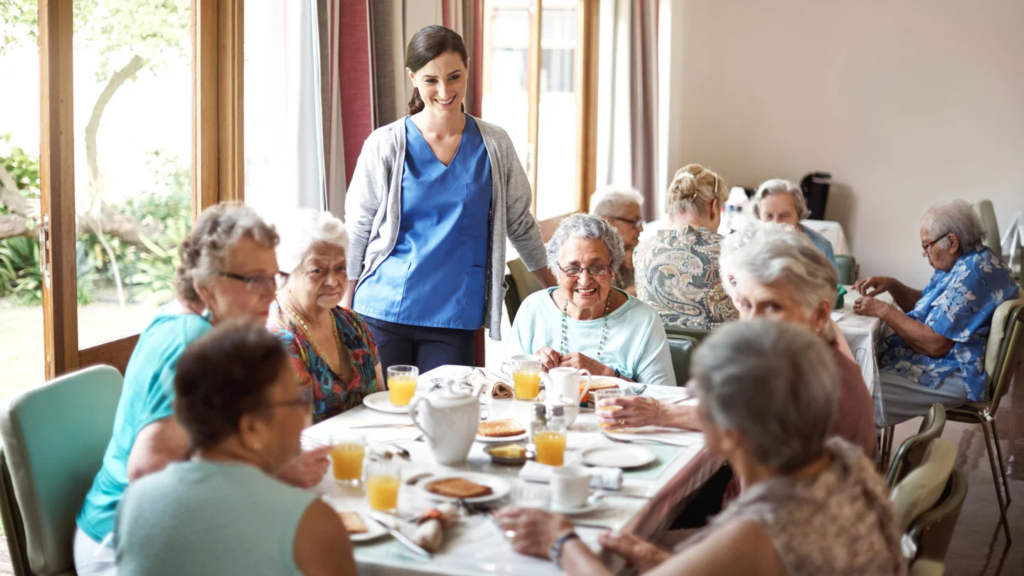 Seniorinnen frühstücken gemeinsam in einer Pflegeeinrichtung, eine junge Mitarbeiterin steht am Tisch.