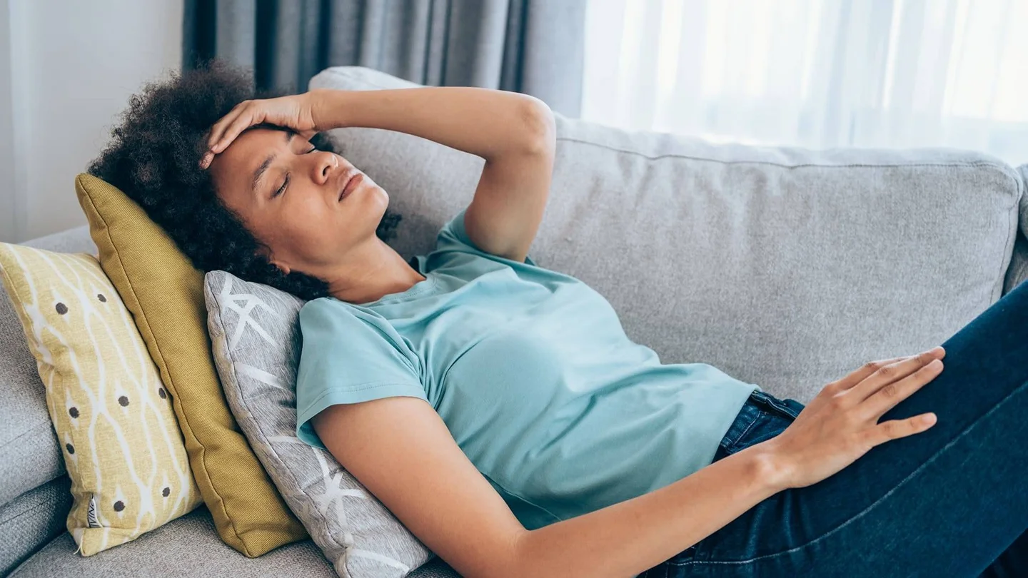 Eine Frau liegt, gestützt auf ein paar Kissen, auf der Couch. Sie greift sich erschöpft an den Kopf.