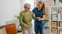 Eine Krankenschwester st&uuml;tzt eine &auml;ltere Frau. Die &auml;ltere Frau benutzt einen Gehstock. Beide l&auml;cheln. 