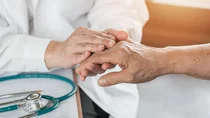 Ein Arzt untersucht die Hand einer gegen&uuml;bersitzenden Person. Auf dem Tisch liegt ein Stethoskop.