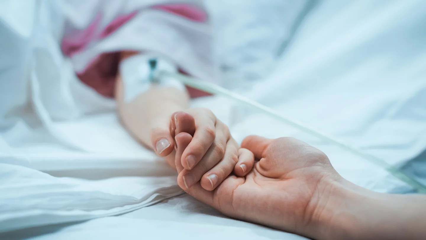 Ein Kind im Krankenhausbett hält die Hand einer erwachsenen Person.