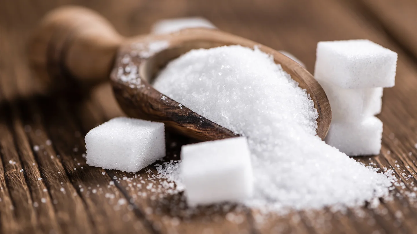 Zucker: Eine Holzschaufel ist mit Zuckerkristallen bef&uuml;llt. Rechts und links neben der Schaufel liegen mehrere Zuckerw&uuml;rfel