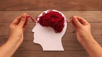 Ein rotes Wollknäuel liegt auf einer weißen Silhouette eines Kopfes. Von unten ragen zwei Hände ins Bild, die die losen Enden des Wollknäuels rechts und links des Kopfes festhalten.