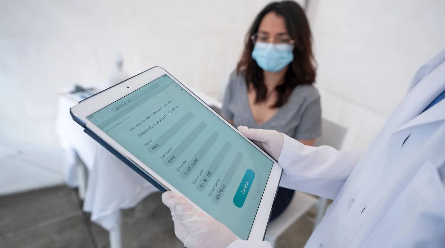 Krankenhaus: Im Vordergrund ein Tablet mit Patientendaten, im Hintergrund eine Patientin.