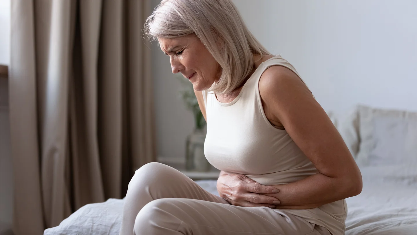 Zwölffingerdarmgeschwür: Eine ältere Frau sitzt auf dem Bett. Sie hält sich den Bauch und zieht die Beine an, ihr Gesicht ist schmerzverzerrt.